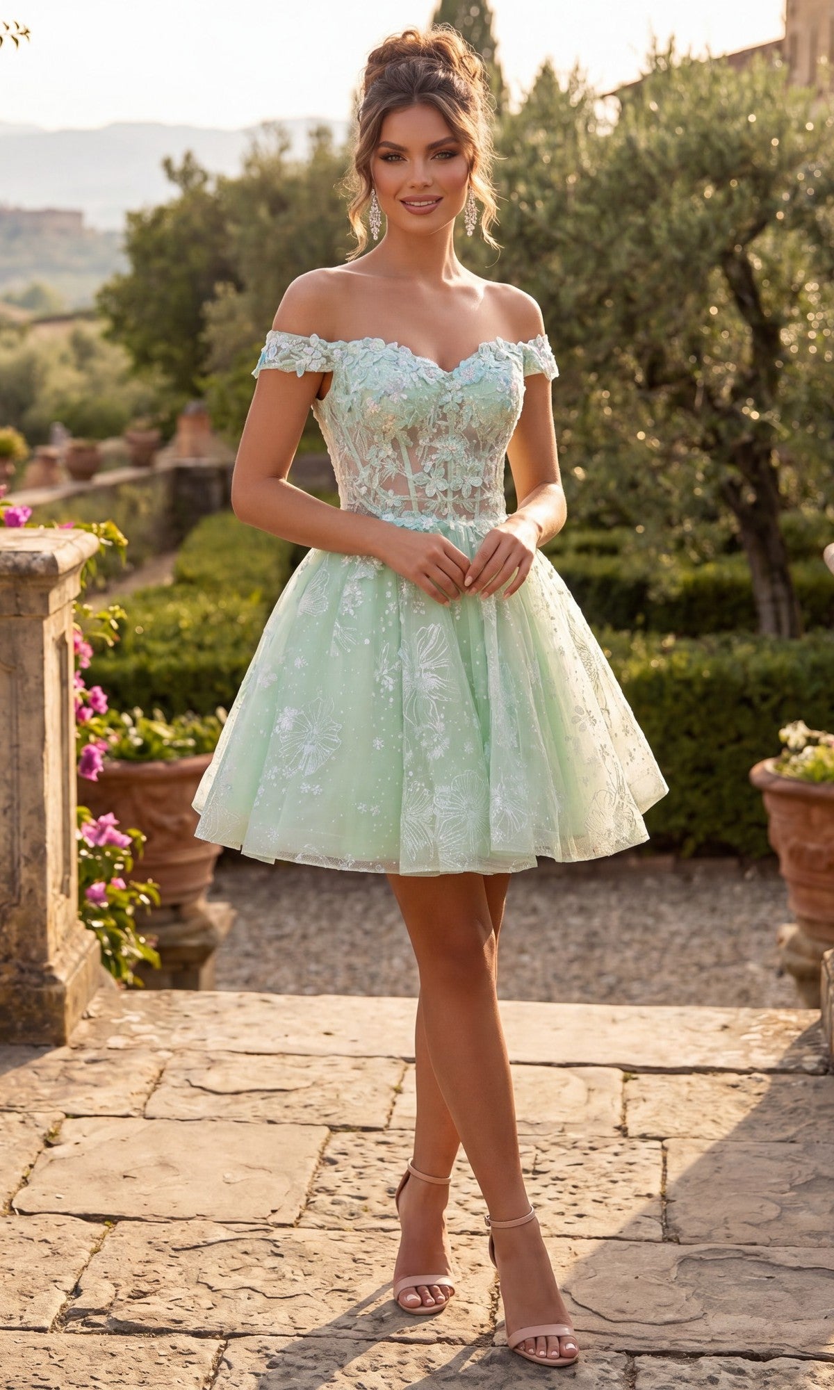 A woman stands outdoors in a garden, smiling at the camera with her hair styled up, wearing the Chicas C377 short homecoming dress in sage—a light green, off-the-shoulder design featuring a lace tulle skirt—paired with nude heels.